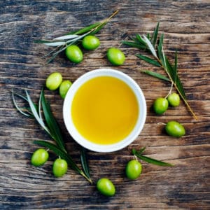 photo of bowl of olive oil on wood table with green olives and leaves around it representing Arbosana Extra Virgin Olive Oil