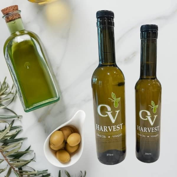 Bottle of olive oil, olive branch, bowl of olives and OV Harvest olive oil bottles representing “Oro Bailen” Picual Extra Virgin Olive Oil