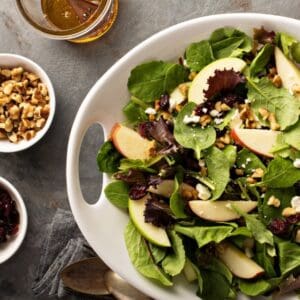 Bowl of salad with leaves, apple, nuts, and dried fruit. Three bowls are sitting next to it on the table with dressing, nuts, and cranberries inside.