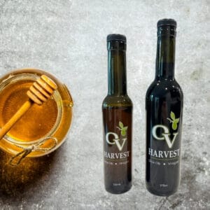 Glass jar of honey with a stone background and OV Harvest vinegar bottles representing Serrano Honey Vinegar