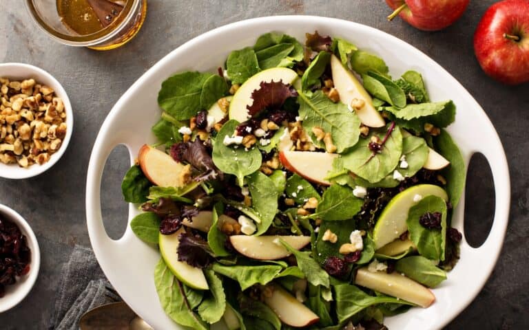 Bowl of salad with leaves, apple, nuts, and dried fruit. Three bowls are sitting next to it on the table with dressing, nuts, and cranberries inside.