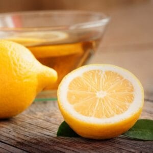 Image of a sliced lemon sitting on a wood table with a bowl of liquid in the background representing Organic Greek Lemon Olive Oil.