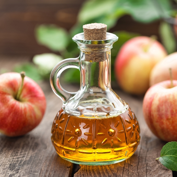 A jar of apple cider vinegar sits on a wood table surrounded by red apples and leaves representing unfiltered organic apple cider vinegar.